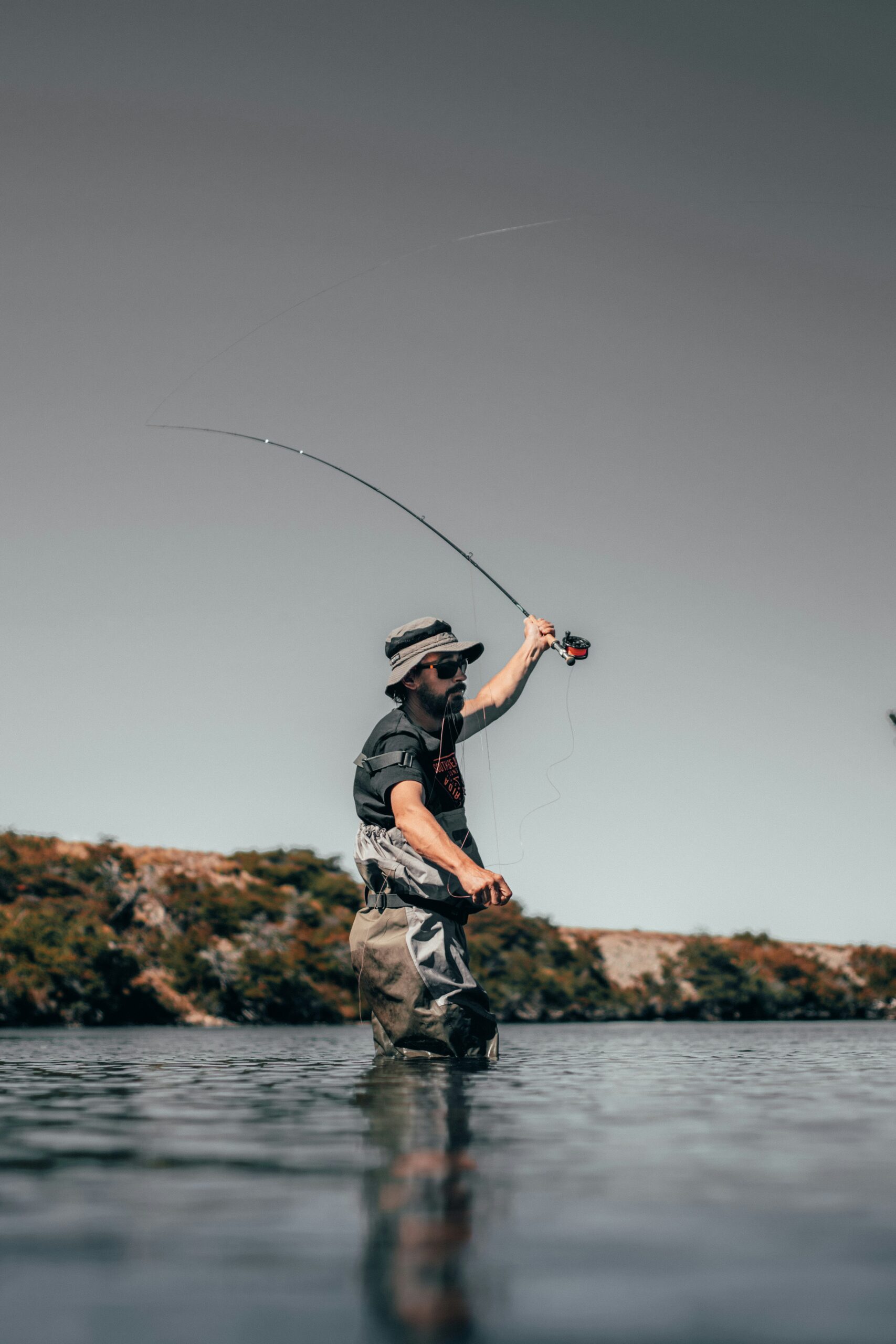 A man wearing waterproof gear stands in the river, casting his fishing rod on a sunny day.


